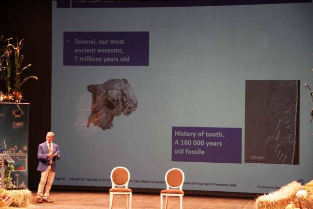 Ein Mann präsentiert auf einer Bühne Informationen über den fossilen Schädel von Toumaï und einen Zahn.