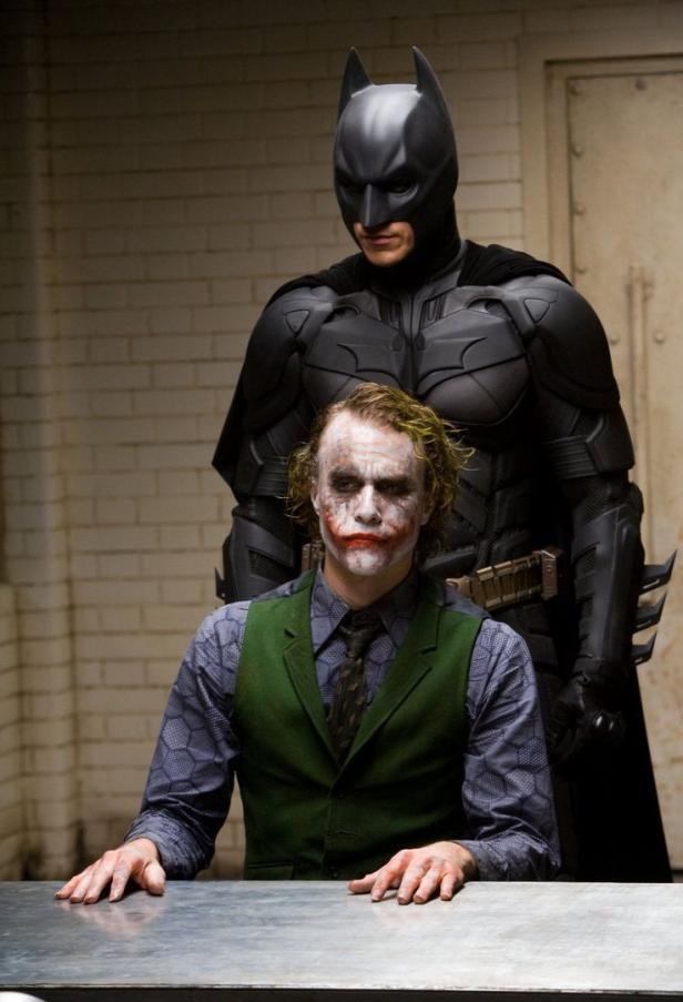 Batman steht hinter dem Joker, der an einem Tisch sitzt.