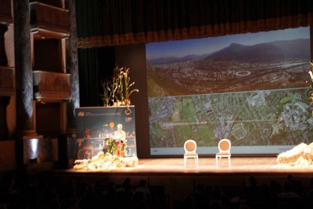 Eine Rednerin auf einer Bühne vor einer Projektion von Luftaufnahmen einer Stadt.