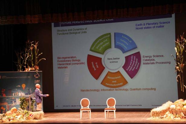 Ein Redner präsentiert eine Folie über neue wissenschaftliche Perspektiven auf einer dekorierten Bühne.