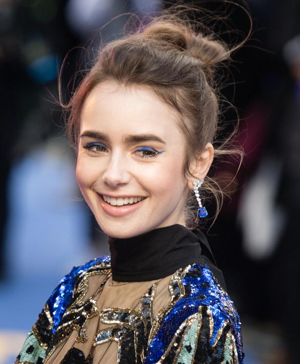Lily Collins lächelt mit blauem Eyeliner und Ohrringen.