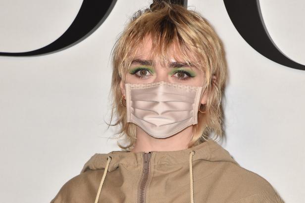 Maisie Williams mit blonden Haaren und grünem Lidschatten trägt eine Maske.