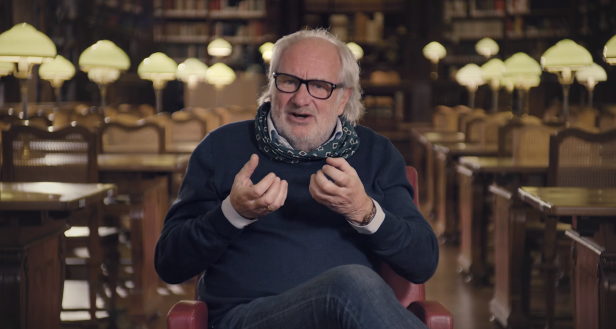 Ein Mann mit Brille und Schal sitzt in einer Bibliothek und gestikuliert.