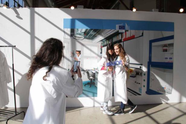 Eine Frau fotografiert zwei junge Frauen in Laborkitteln vor einem Wandbild mit Labormotiven.