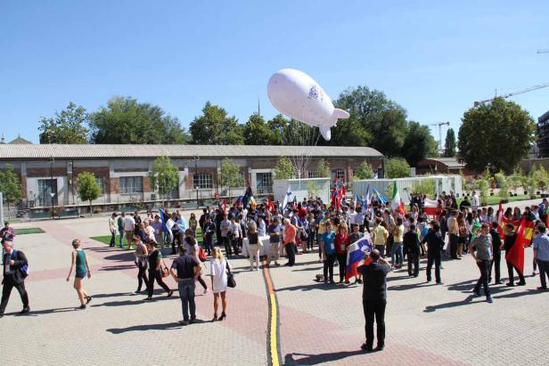 Eine Menschenmenge versammelt sich im Freien mit Flaggen verschiedener Länder und einem weißen Luftschiff.