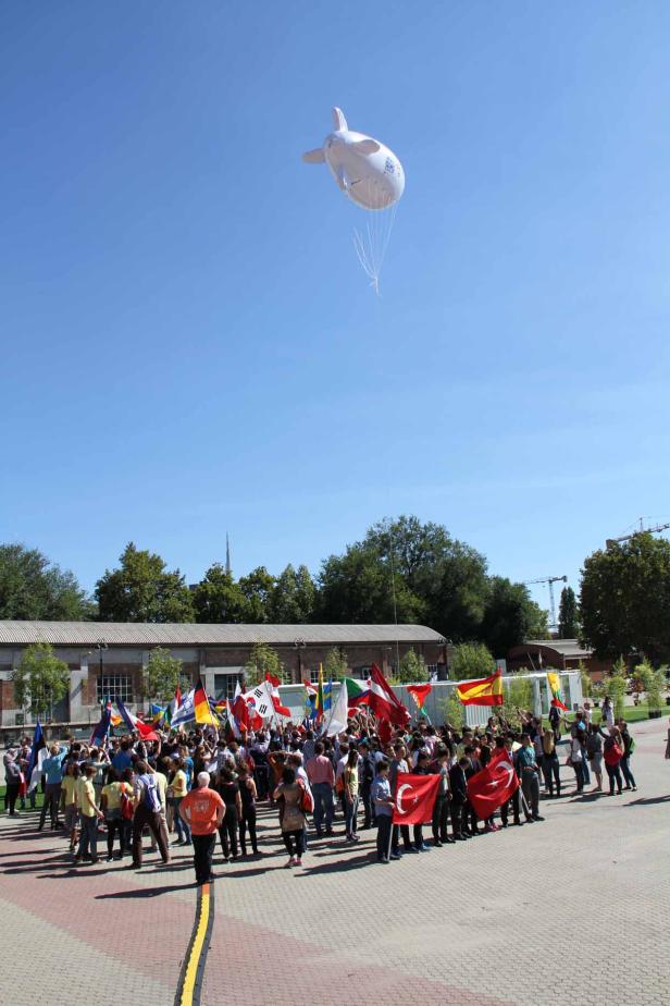 Eine Menschenmenge hält Flaggen verschiedener Länder unter einem weißen Luftschiff.