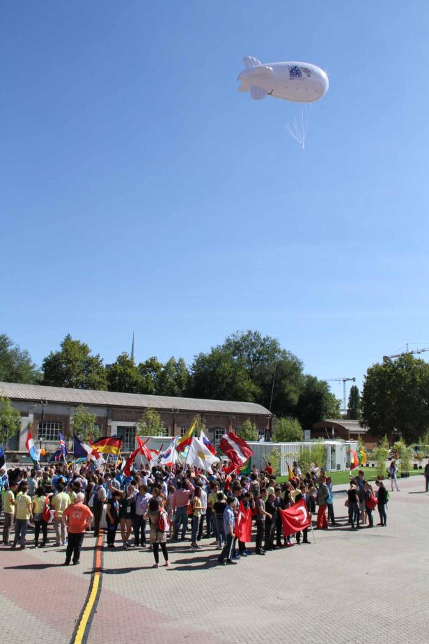 Eine Menschenmenge hält verschiedene Nationalflaggen unter einem weißen Luftschiff.