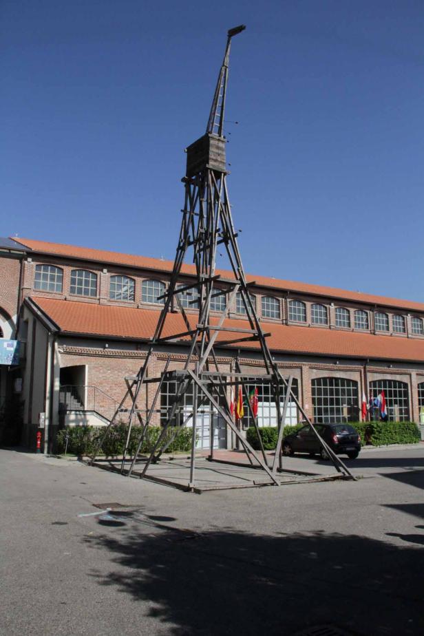 Eine hölzerne Kran-Skulptur steht vor einem Backsteingebäude unter blauem Himmel.