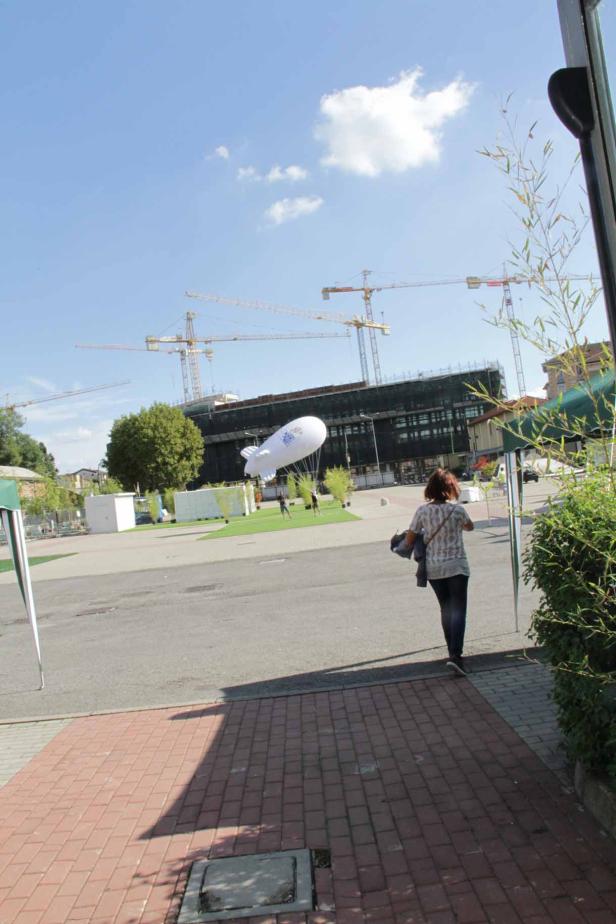 Eine Frau geht auf einen Platz zu, im Hintergrund ein Luftschiff und mehrere Baukräne.