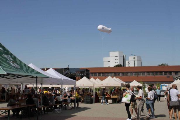 Ein belebter Marktplatz mit Zelten und einem kleinen Luftschiff am Himmel.