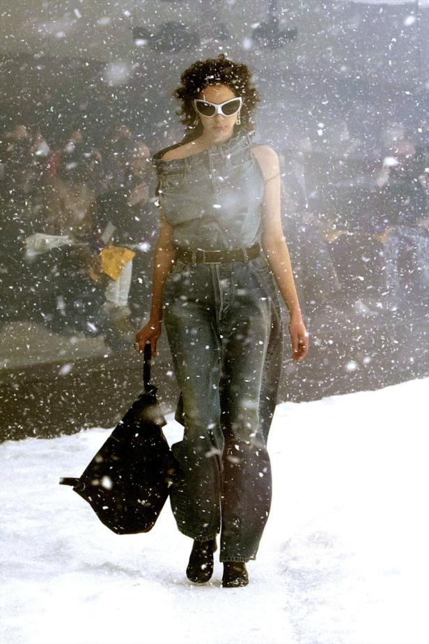 Eine Frau mit Sonnenbrille und Jeanskleidung geht durch den Schnee.