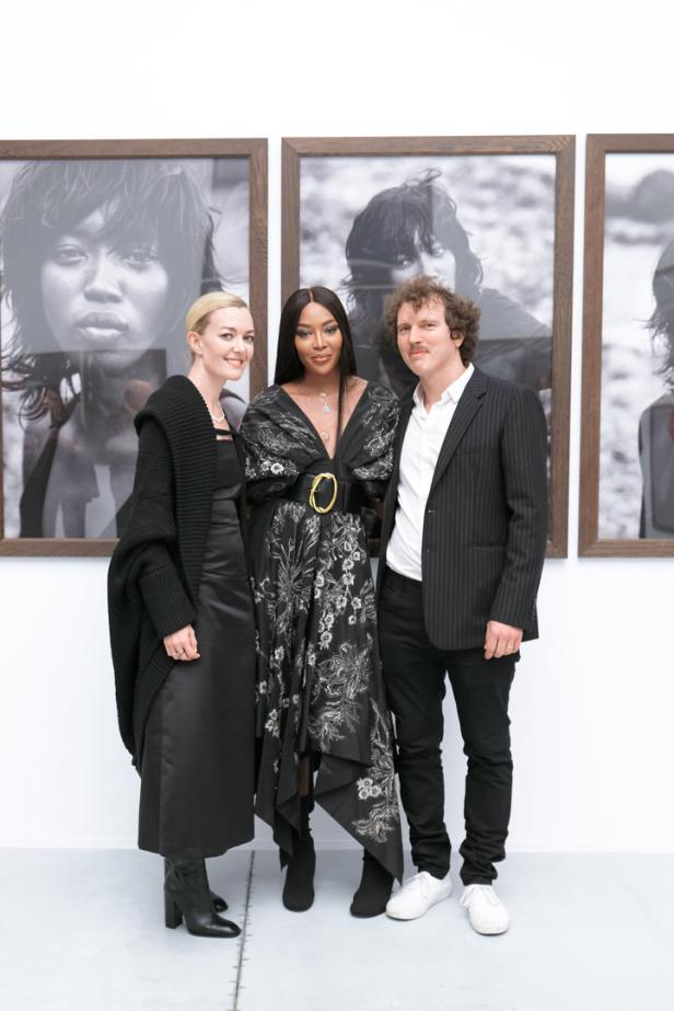 Naomi Campbell posiert mit zwei weiteren Personen vor Porträts.