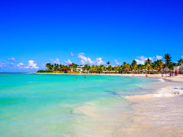 Ein tropischer Strand mit türkisfarbenem Wasser, weißem Sand und Palmen unter blauem Himmel.