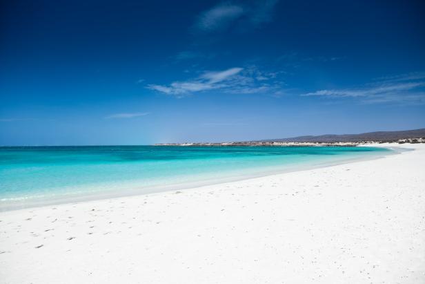 Ein strahlend weißer Sandstrand trifft auf türkisfarbenes Wasser unter blauem Himmel.