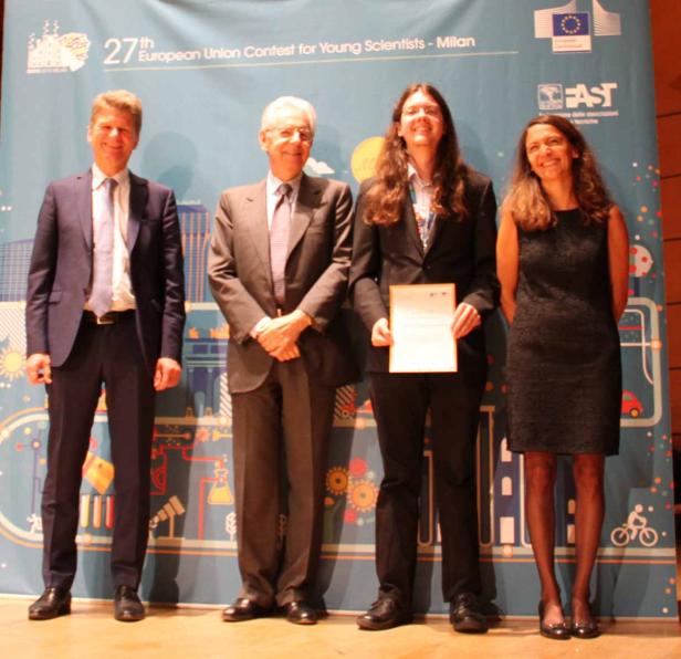 Vier Personen posieren vor einem Banner des 27. Europäischen Wettbewerbs für junge Wissenschaftler in Mailand.