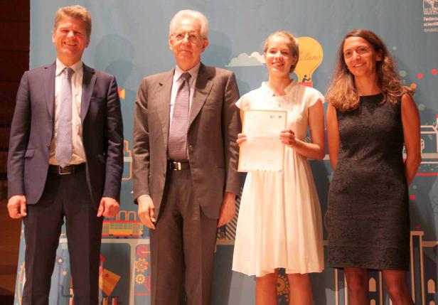 Eine junge Frau hält eine Urkunde zwischen drei Personen auf einer Bühne.