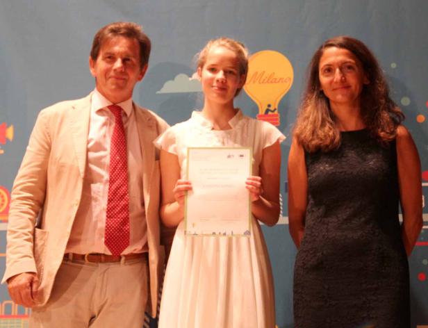 Ein junges Mädchen hält eine Urkunde zwischen zwei Erwachsenen vor einem farbenfrohen Hintergrund.