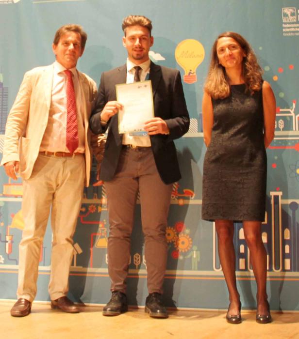 Drei Personen posieren vor einem Hintergrund mit wissenschaftlichen Motiven und dem Schriftzug „Milano“.