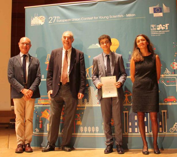 Vier Personen posieren vor einem Banner des 27. Europäischen Wettbewerbs für junge Wissenschaftler in Mailand.