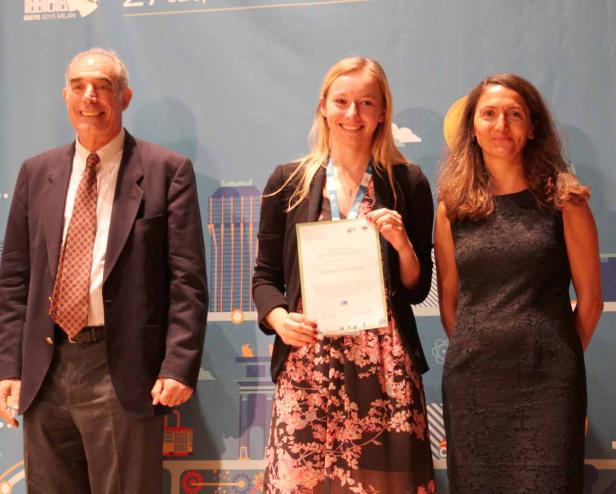 Eine junge Frau hält eine Urkunde zwischen zwei Personen auf einer Bühne.