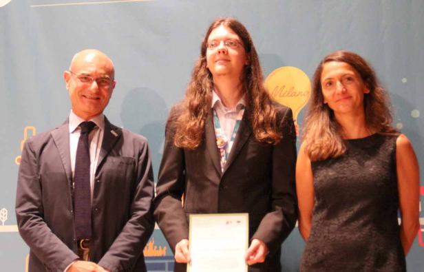 Drei Personen posieren vor einem Hintergrund mit dem Schriftzug „Milano“.