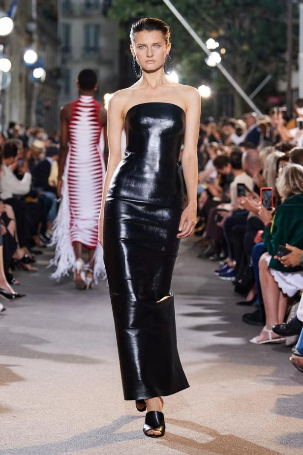 Ein Model präsentiert ein trägerloses, glänzendes, schwarzes Kleid auf einem Laufsteg.