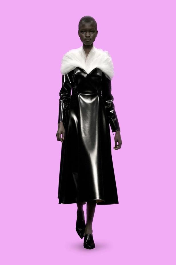 Eine Frau mit dunkler Haut trägt ein schwarzes Lackkleid mit weißem Kragen vor einem violetten Hintergrund.