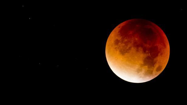 Eine totale Mondfinsternis leuchtet rot und orange vor einem schwarzen Nachthimmel.