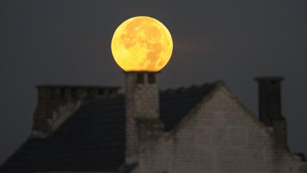 Ein gelber Vollmond geht über einem Hausdach auf.