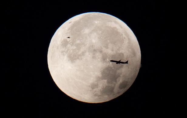Ein Flugzeug und ein kleineres Flugobjekt fliegen vor dem Hintergrund des Vollmonds.