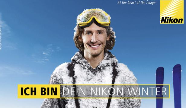 Ein lächelnder Mann mit Skibrille und Schnee auf der Kleidung vor blauem Himmel.