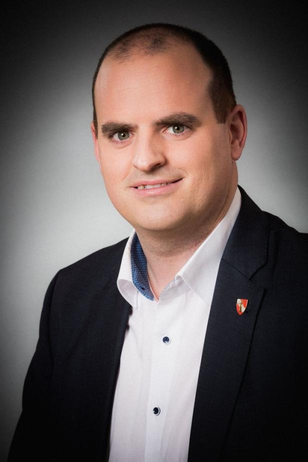 Ein Mann im Anzug mit einem Wappen am Revers lächelt in die Kamera.