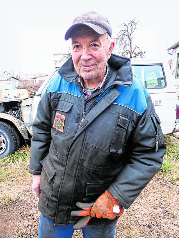 Ein älterer Mann mit Mütze und Handschuhen steht vor einem beschädigten Auto.