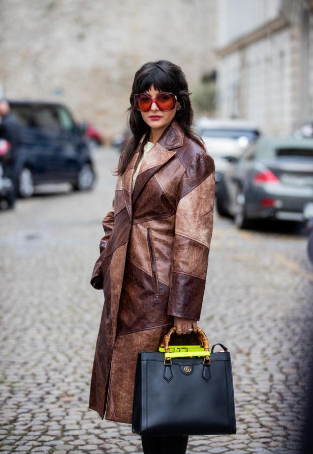 Eine Frau mit Sonnenbrille und braunem Ledermantel hält eine schwarze Gucci-Tasche.