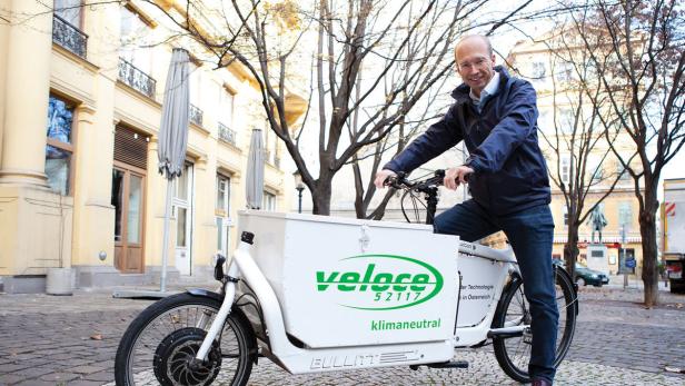 Firmenchef Brandstätter Ein Mann posiert auf einem weißen Lastenfahrrad der Marke „Veloce“ in einer städtischen Umgebung.