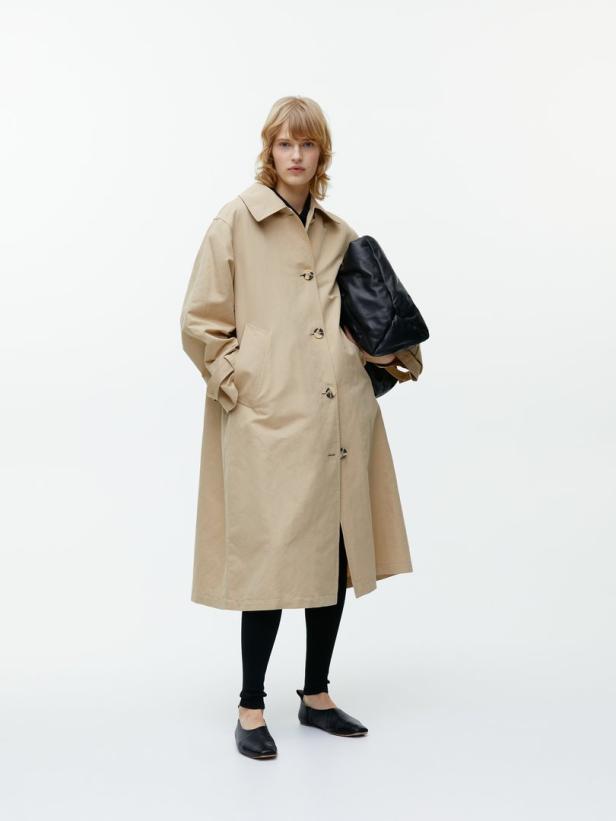 Eine Frau mit blonden Haaren trägt einen beigen Trenchcoat, Leggings und eine schwarze Tasche.