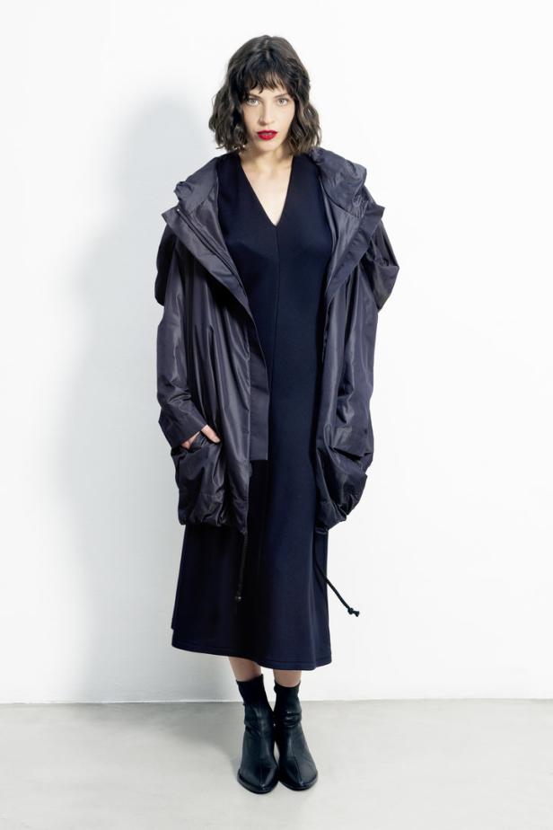 Eine Frau mit rotem Lippenstift trägt ein schwarzes Kleid und eine übergroße Jacke mit Kapuze.