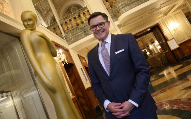 Ein lächelnder Mann im Anzug steht neben einer goldenen Statue in einem eleganten Gebäude.