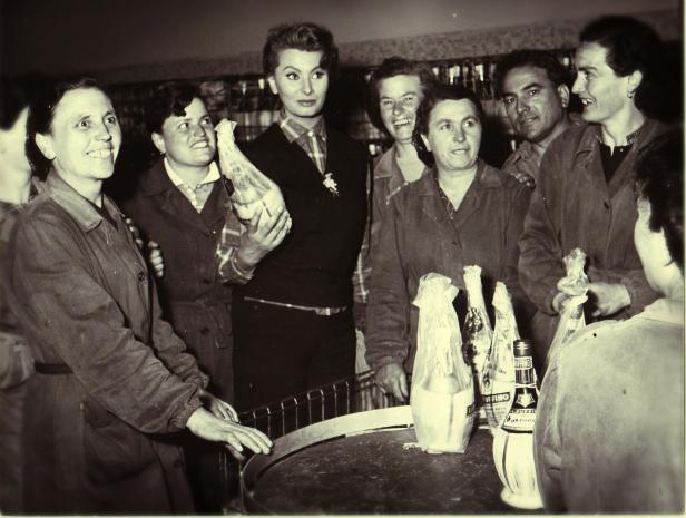Sophia Loren umgeben von Arbeiterinnen und Arbeitern mit Flaschen auf einem Tisch.