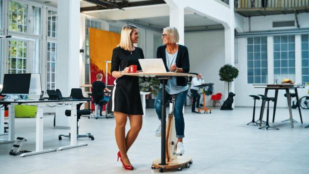 Zwei Frauen unterhalten sich an einem Stehtisch mit integriertem Fahrrad in einem Büro.