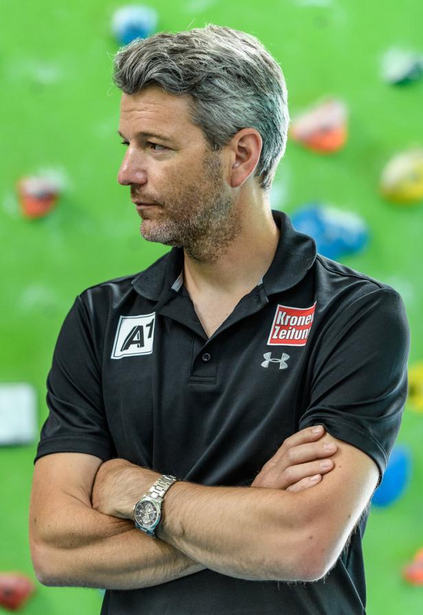 Ein Mann mit verschränkten Armen vor einer grünen Wand mit Klettergriffen.