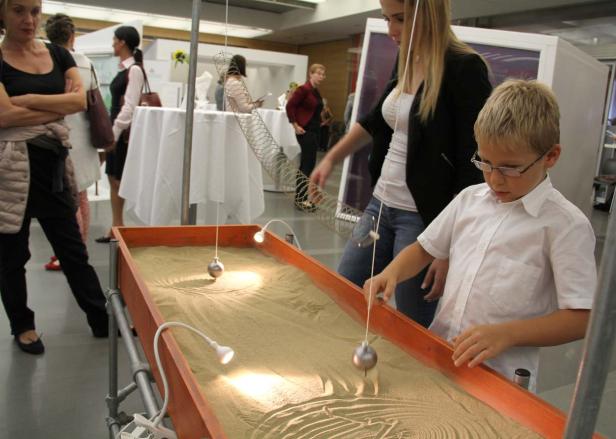 Ein Junge mit Brille beobachtet ein Pendel, das Muster in Sand zeichnet.