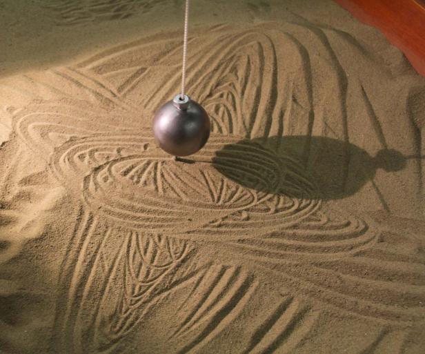 Ein Pendel hängt über einem Sandkasten und erzeugt Muster.