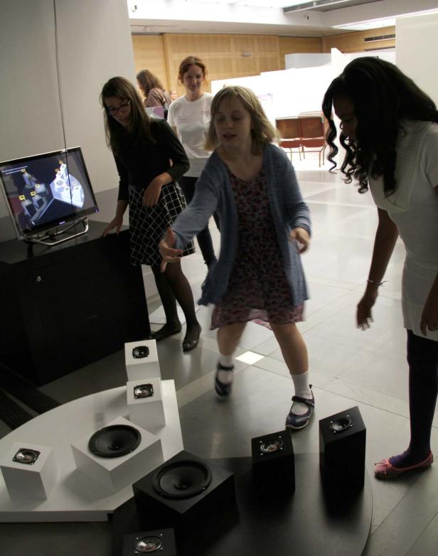 Drei junge Frauen interagieren mit einer Installation aus Lautsprechern und einem Bildschirm.
