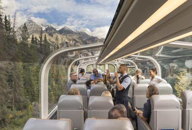 Ein Zug mit Panoramablick fährt durch eine Landschaft mit Bergen und Wäldern, während ein Mann eine Tour gibt.