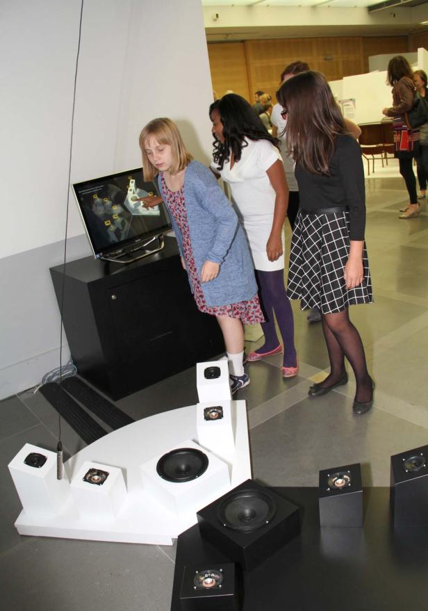 Drei Mädchen betrachten eine interaktive Installation mit Lautsprechern und einem Bildschirm.