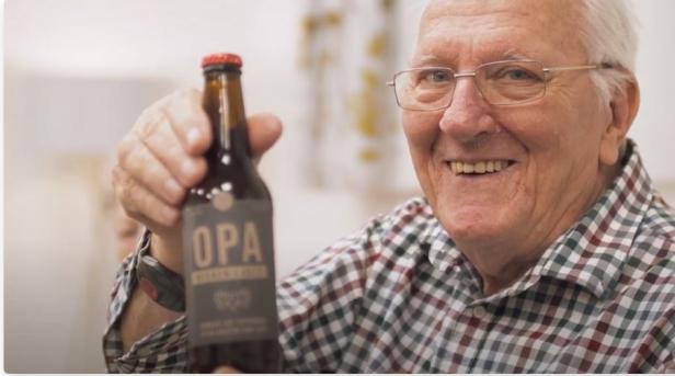 Ein älterer Mann lächelt und hält eine Flasche „Opa“-Bier hoch.