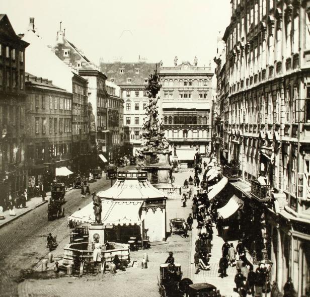 Historische Aufnahme einer belebten Straße in Wien mit der Pestsäule im Zentrum.