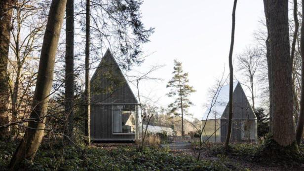 Zwei moderne, dreieckige Hütten stehen in einem lichten Wald.