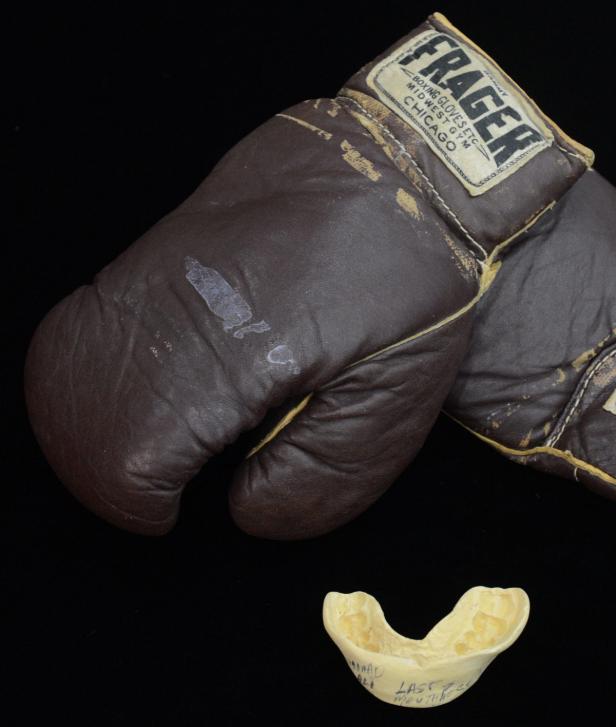 Ein Paar braune Frager-Boxhandschuhe und ein Mundschutz liegen auf einem schwarzen Hintergrund.